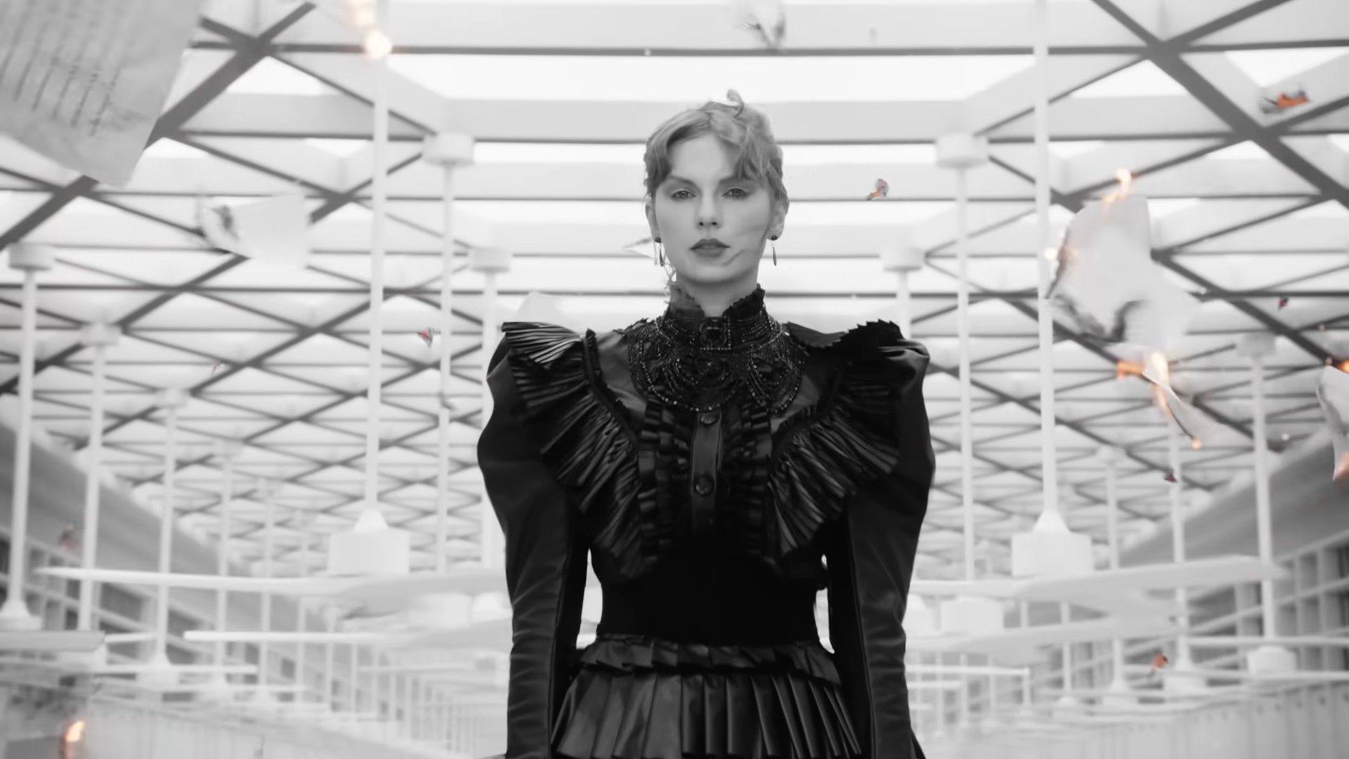 Taylor Swift in a black dress standing in the middle of a room with papers flying around it